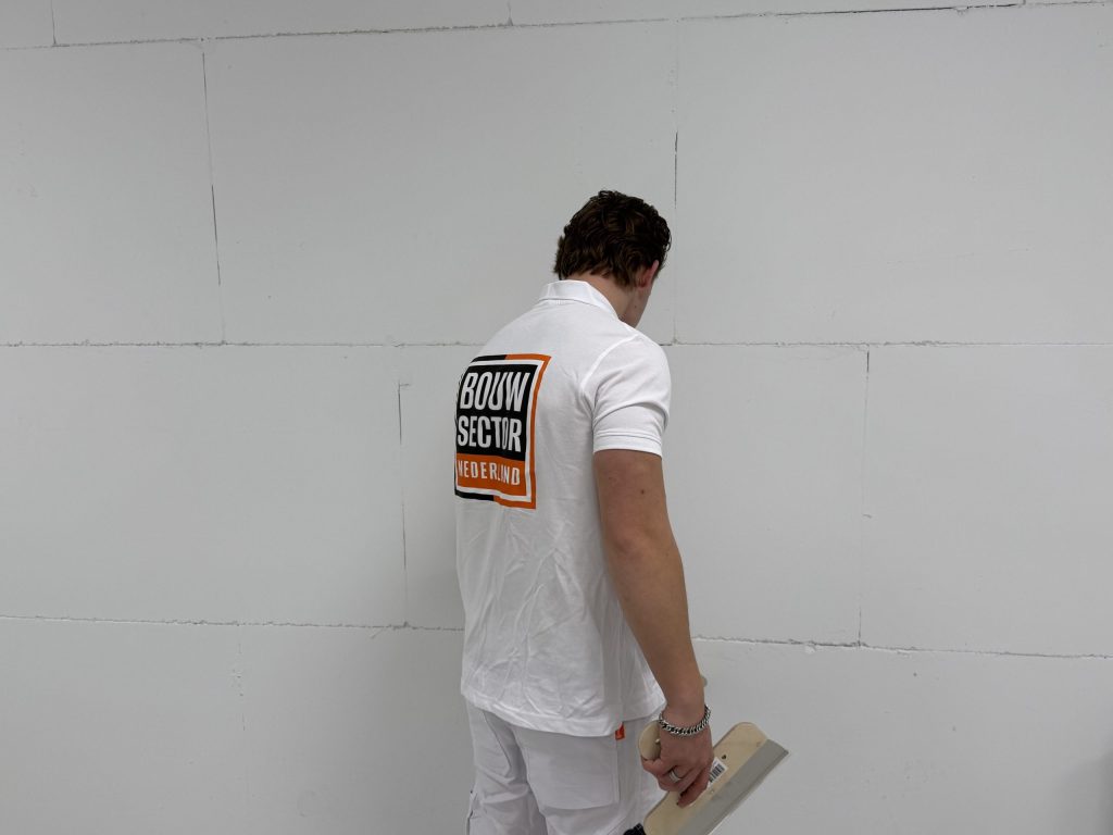 Stukadoor in wit poloshirt met het logo van Bouwsector Nederland bekijkt een kale wand, spaan in de hand