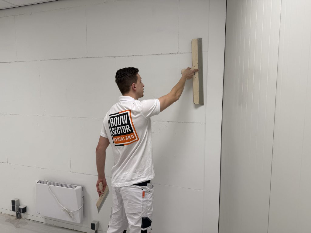 Stukadoor strijkt met een brede spaan over een wand boven een elektrische kachel