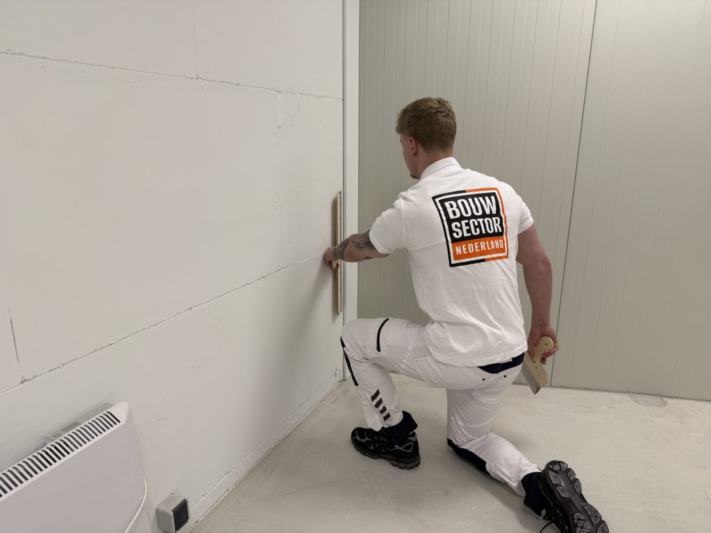 Knielende stukadoor in wit werktenue van Bouwsector Nederland trekt een rei langs de onderkant van een wand